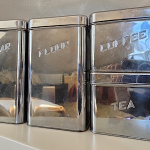 Peut inclure: Un ensemble de quatre boîtes de cuisine rectangulaires en chrome poli avec des coins arrondis et des couvercles ajustés. Les boîtes sont gravées avec les mots SUGAR, FLOUR, COFFEE et TEA dans une police sans empattement sur la face avant.