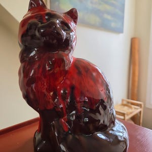 May include: A ceramic cat figurine featuring a glossy finish with a marbled colour pattern of deep black and vibrant red. The cat is depicted in a seated position with its head tilted upward, showcasing a smooth, reflective surface throughout.