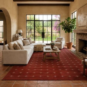 Puede incluir: Una alfombra rectangular grande de color rojo terracota intenso con un patrón repetitivo de pequeñas cruces blancas. La alfombra está colocada sobre un suelo de baldosas de terracota en una sala de estar con un sofá seccional beige y una mesa.