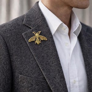Könnte beinhalten: Eine goldfarbene Bienenbrosche, die an einem grauen Blazer befestigt ist. Die Bienenbrosche ist mit Flügeln und Körpersegmenten detailliert. Der Blazer wird über einem weißen Hemd getragen.