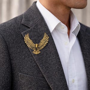 Może przedstawiać: Złota broszka w kształcie orła przypięta do szarej marynarki. Broszka, ze szczegółowymi rozłożonymi skrzydłami, jest noszona na białej koszuli z kołnierzykiem. Marynarka jest w ciemnoszarym kolorze.