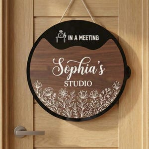 Puede incluir: Cartel de puerta redondo de madera con el texto "Sophia's STUDIO" en escritura blanca y un diseño floral en la parte inferior. La sección superior es negra con las palabras "IN A MEETING" y un pequeño gráfico. El letrero cuelga de una cuerda natural.