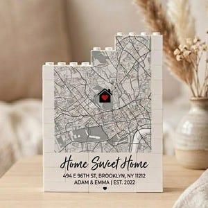 Puede incluir: Una pieza decorativa blanca en forma de bloque con un diseño de mapa y un icono de casa en forma de corazón rojo. El texto "Home Sweet Home" se muestra debajo del mapa, junto con una dirección y el texto "ADAM & EMMA | EST. 2022".