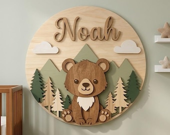 May include: A wooden wall decoration featuring a bear, mountains, trees, and clouds. The name "Noah" is displayed in a cursive font. The bear is brown with a white chest. The mountains are green, and the trees are various shades of green and brown.