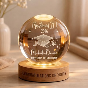 May include: A clear glass sphere with the text "Mastered It! 2026" and a graduation cap and diploma design. The sphere sits on a wooden base with the words "CONGRATULATIONS ON YOURS".