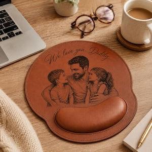 Peut inclure: Un tapis de souris en cuir marron avec repose-poignet. Le tapis présente un portrait de famille et le texte "We love you, Daddy". L'image montre un père et deux enfants. Le tapis est sur un bureau en bois.
