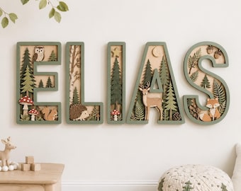 May include: A wall-mounted wooden name sign spelling ELIAS in large, sage green letters. Each letter features a layered, 3D forest scene with woodland animals including an owl, hedgehog, deer, and fox, alongside pine trees and mushrooms.