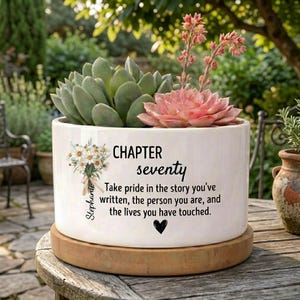 Könnte beinhalten: Ein weißer Keramik-Blumentopf mit dem Text "CHAPTER seventy" und einer floralen Bouquet-Illustration. Der Topf enthält grüne und rosa Sukkulenten. Der Topf steht auf einem Holzsockel, mit einem Gartenhintergrund.