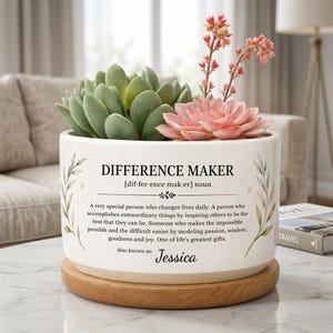 Puede incluir: Una maceta de cerámica blanca con el texto "DIFFERENCE MAKER" y una definición. La maceta contiene una suculenta con hojas verdes y rosas. La maceta se asienta sobre una base de madera. El nombre "Jessica" también está en la maceta.