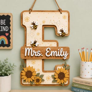 May include: A wooden wall hanging in the shape of the letter E, featuring a cream-colored background with yellow flower patterns and three small bee cutouts. The name Mrs. Emily is written in white script, and two large sunflowers with green leaves.