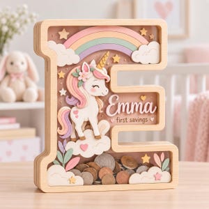 Può includere: Un salvadanaio in legno a forma di lettera E con un pannello frontale trasparente. All'interno, un unicorno bianco con criniera rosa e viola si trova su nuvole sotto un arcobaleno. Il testo Emma e first savings è stampato sul retro. Monete riempiono la base.