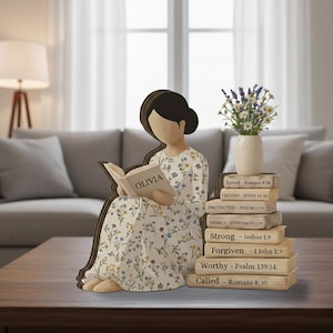 Puede incluir: Una figura de madera de una mujer leyendo un libro con el nombre "OLIVIA" en la portada. La mujer lleva un vestido floral. A su lado hay una pila de libros con palabras inspiradoras y versículos bíblicos. Un jarrón con flores se encuentra encima de los libros.