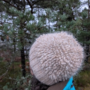 May include: A beige knitted hat with a fluffy, textured top. The hat is worn by a person wearing a blue jacket, with a forest background.
