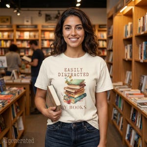 以下が含まれることがあります： クリーム色の半袖Tシャツで、本の積み重ねのグラフィックと「EASILY DISTRACTED BY BOOKS」という茶色の文字がプリントされています。濃い青のデニムジーンズと合わせて着用し、片手に本を一冊持っています。