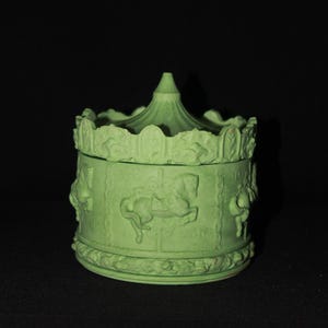 Op de afbeelding: Een lichtgroene sieradendoos met carrouselthema. De doos heeft reliëf carrouselpaarden en ruiters rondom de zijkanten, met een decoratieve boven- en onderkant. Het deksel heeft een kleine, conische top. De doos is gemaakt van keramiek of een vergelijkbaar materiaal.