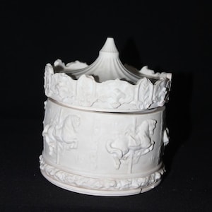 Op de afbeelding: Witte keramische sieradendoos met een carrouselthema. De doos heeft een deksel met een tentachtig ontwerp en een basis versierd met carrouselpaarden in reliëf. Een decoratief item.
