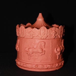 Op de afbeelding: Een koraalkleurige sieradendoos met carrouselthema. De doos heeft reliëf carrouselpaarden en ruiters aan de zijkanten, met een geschulpte bovenkant en een conisch dak. De doos is gemaakt van een stevig materiaal en heeft een matte afwerking.