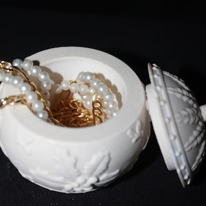 Könnte beinhalten: Eine weiße, runde Schmuckschatulle mit offenem Deckel, die Perlenketten und goldene Ketten zeigt. Die Schatulle hat ein florales Design und ist vor einem dunklen Hintergrund platziert.