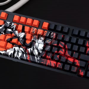 Pode incluir: Um teclado mecânico preto com teclas laranja e pretas. Algumas teclas apresentam um design gráfico em vermelho, preto e branco. O teclado foi projetado para jogadores e entusiastas de computadores.