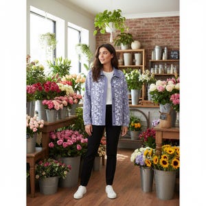 Pode incluir: Uma mulher usa uma jaqueta lilás clara com um desenho floral branco, calças pretas e tênis brancos em uma floricultura. A loja está cheia de flores em baldes de metal e prateleiras de madeira.