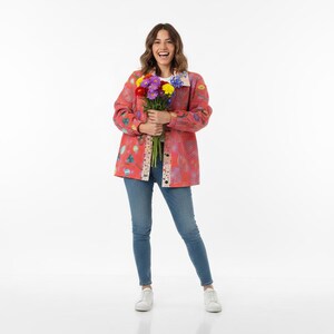 May include: A coral-coloured, button-down jacket with a colourful floral pattern and a patterned trim. The jacket is worn with blue jeans and white trainers. A bouquet of colourful flowers is held in front.