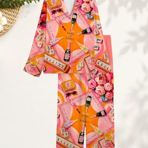May include: A long-sleeve button-down pajama top and matching pants set in a vibrant pink and orange color palette. The fabric features a repeating graphic print of champagne bottles, mahjong tiles, pink peonies, and the text BAMS & BUBBLY.