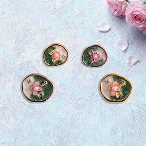 May include: Four floral-themed earrings with pink and white enamel flowers, green leaves, and a pearl center. The earrings have gold or silver-colored metal edging and small crystal accents. Pink roses and petals are in the background.