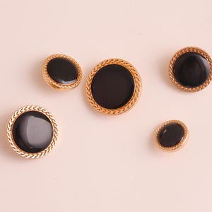 May include: Five gold-toned buttons with black centers. Two are oval-shaped, and three are round. The buttons have a textured, rope-like border. The buttons are arranged on a light pink background.