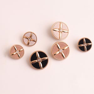 May include: A collection of decorative buttons in various sizes and colors. The buttons feature a central jewel and a cross-shaped design, with gold-colored rims. Colors include black, pink, and beige.