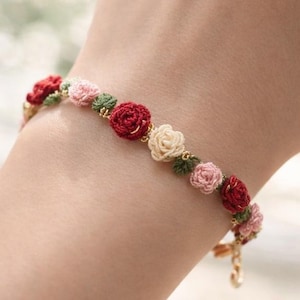 Könnte beinhalten: Ein zartes Armband mit kleinen, gehäkelten Rosen in Rot-, Rosa- und Cremetönen, die mit grünen Blättern durchsetzt sind. Das goldfarbene Armband wird am Handgelenk getragen.