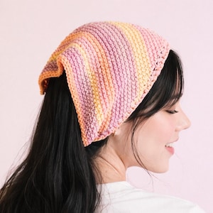 May include: A knitted bandana in shades of pink, orange, and yellow. The head covering is square-shaped and appears to be handmade. The bandana is worn on the head.
