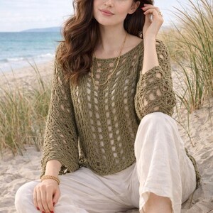 May include: An olive green crochet sweater with long sleeves and a round neckline. The sweater has an open, airy design. Paired with white linen trousers, the outfit is accessorized with gold jewellery. The background features a beach scene with sand and ocean.