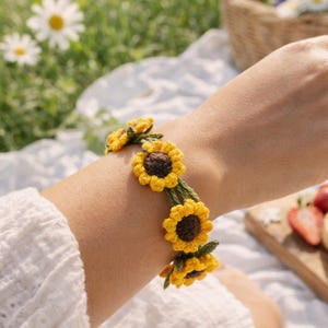 Puede incluir: Una pulsera de ganchillo con girasoles amarillos, centros marrones y tallos verdes. La pulsera se lleva en la muñeca, con un entorno de picnic en el fondo. Un accesorio encantador.