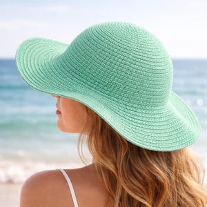 May include: A mint green sun hat with a wide brim. The hat is made of woven material and has a rounded crown. The image shows the hat from a side view, with the ocean in the background. The hat is a stylish accessory for sun protection.