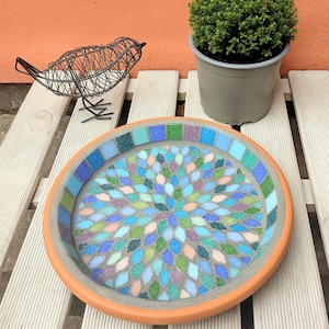 May include: A round, terracotta bird bath with a mosaic design in shades of blue, green, and purple. The bird bath is sitting on a white wooden surface.