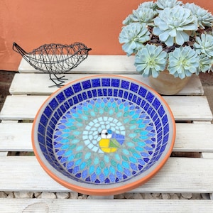 May include: A round, mosaic bird bath with a blue and turquoise design, featuring a central bird motif. The bird bath has a terracotta-colored rim and sits on a white wooden surface. A wire bird sculpture and a potted succulent plant are also present.