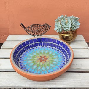 May include: A round, terracotta bird bath with a mosaic tile design. The mosaic features a blue border and a central floral pattern in shades of blue, green, and orange. A wire bird sculpture and a potted succulent are also present.