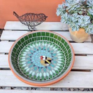May include: A mosaic bird bath with a detailed design featuring a bird in the center, surrounded by a pattern of green, blue, and white tiles. The bird bath has an orange rim and sits on a white wooden surface.