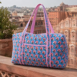 Puede incluir: Un bolso de viaje acolchado con base de tela azul y un estampado floral repetitivo en rosa y verde. El bolso incluye dos asas largas y detalles de ribete en tela a rayas rosas y blancas, con un bolsillo frontal de forma rectangular.