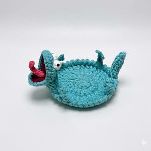 Könnte beinhalten: Ein handgefertigtes, türkisfarbenes Häkeltier mit weit geöffnetem Maul und rosa Zunge. Das skurrile Design zeigt weiße Augen und kleine Flossen. Das Tier liegt auf dem Rücken und präsentiert sein verspieltes, cartoonartiges Aussehen.