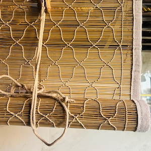 Op de afbeelding: Een natuurlijke bamboe rolgordijn met een geweven ontwerp. Het rolgordijn is gemaakt van dunne, lichtbruine bamboe latten, verbonden door een beige geweven patroon. Een beige stoffen rand omlijst de boven- en zijkanten. Een touw is bevestigd om het rolgordijn op en neer te halen.