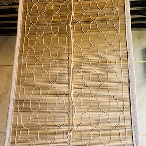 Op de afbeelding: Een rechthoekige bamboe raambekleding met een geweven patroon. De bekleding is omlijst met een lichtbeige stof en heeft een decoratief touw om omhoog en omlaag te trekken. De bamboe latten hebben een natuurlijke lichtbruine kleur.