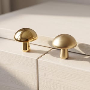 May include: Two mushroom-shaped cabinet knobs made of metal. One knob features a highly reflective, polished gold finish, while the other knob has a muted, brushed gold finish. Both knobs have a rounded cap and a cylindrical base.
