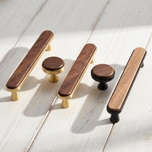 May include: A collection of cabinet hardware including three rectangular drawer pulls and two round knobs. The hardware features a combination of dark brown wood grain inserts set into either brushed gold or matte black metal bases.