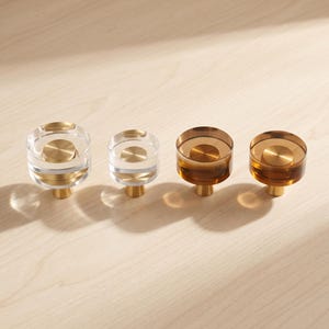 Puede incluir: Cuatro pomos circulares para armarios dispuestos en fila. Dos pomos están hechos de material transparente incoloro con un disco metálico central dorado, y dos pomos son de material transparente color ámbar con un disco metálico central dorado.