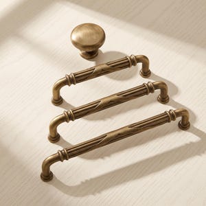 May include: A set of antique brass hardware including one round cabinet knob and three elongated drawer pulls. Each pull features a central fluted design with decorative accents and curved ends, showcasing a brushed metallic finish throughout.