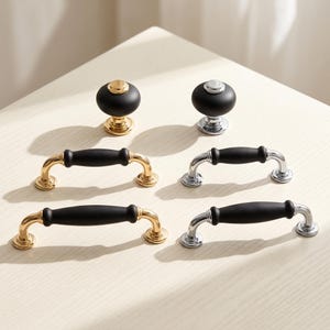 May include: A collection of cabinet hardware including two round knobs and four arched drawer pulls. The hardware features a matte black finish on the central sections, paired with either polished gold-tone or polished silver-tone metal accents.