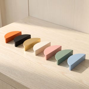 Könnte beinhalten: Ein Set aus sieben halbkreisförmigen Schrankgriffen, die in einer diagonalen Linie angeordnet sind. Die Griffe haben eine matte Oberfläche in Orange, Schwarz, Gold, Creme, Rosa, Salbeigrün und Hellblau mit flacher Oberkante und gewölbter Front.