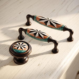 May include: Two metal drawer pulls and one round cabinet knob featuring a dark bronze finish. Each piece has a central ceramic inlay with a black and white starburst pattern surrounded by a tan border and a textured teal green outer edge.