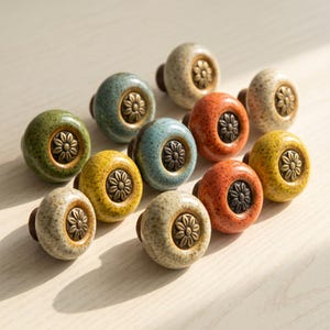 May include: A collection of eleven round ceramic cabinet knobs in various speckled colors including green, blue, yellow, orange, and cream. Each knob features a central bronze-toned metal inlay with a raised floral daisy pattern.